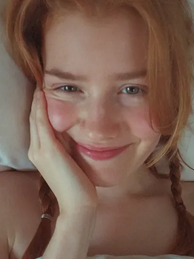 Selfie in bed: red braids, rosy cheeks, hand on her cheek.
